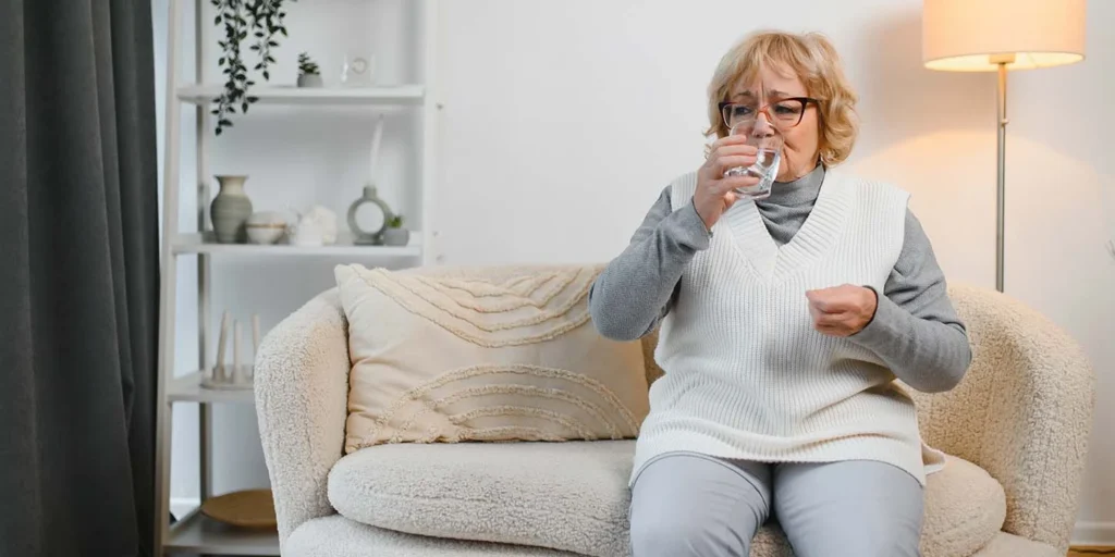 Mulher idosa sentada em um sofá enquanto bebe um copo d'água e segura uma pílula, referenciando o Parkinsonismo Induzido por Medicamentos (DIP).