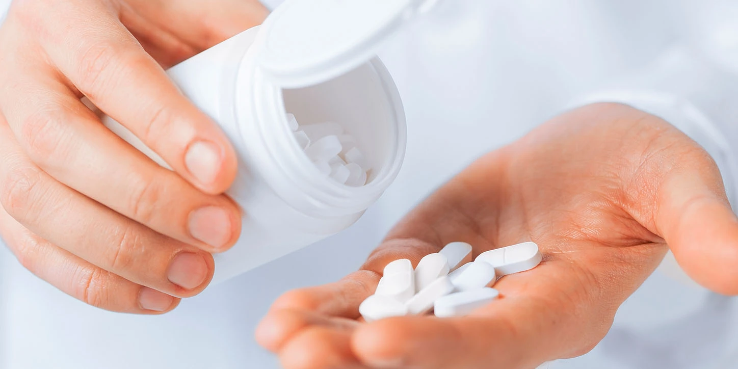 Mãos de uma pessoa derramando várias pílulas brancas de um frasco branco aberto na palma da mão, referenciando o Parkinsonismo Induzido por Medicamentos (DIP)