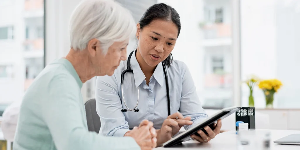 Médica especialista conversando com paciente idosa enquanto aponta dados em um tablet, explicando o plano de cuidado para a Demência com Corpos de Lewy.