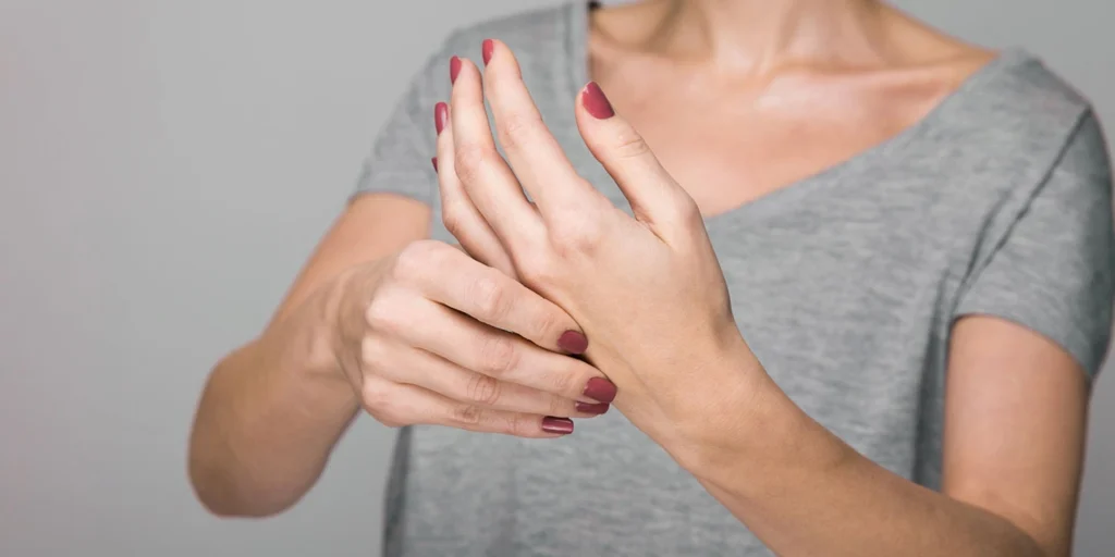 Mulher segurando e massageando a própria mão, sugerindo rigidez ou perda de controle motor, sintomas comuns na Degeneração Corticobasal.